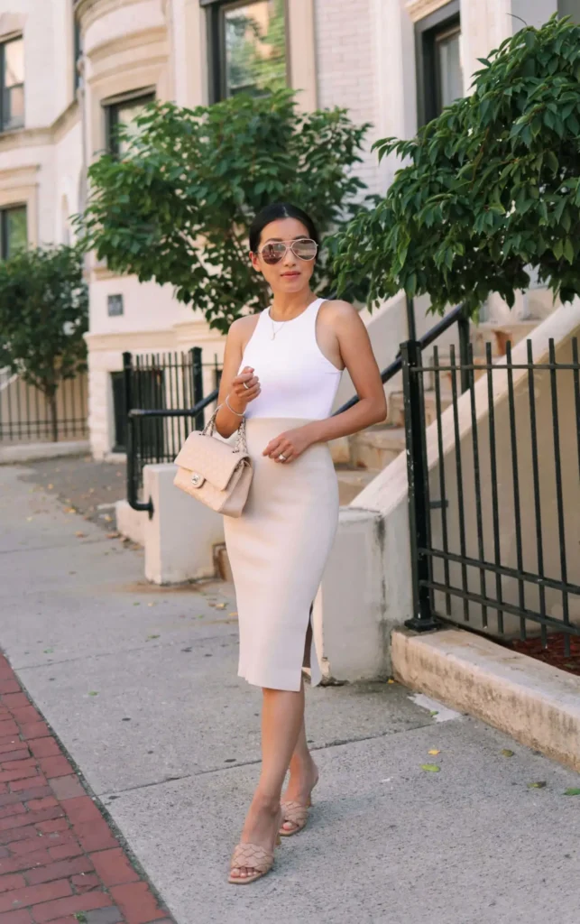 Petite woman in a white sleeveless top tucked into a cream midi pencil skirt with a front slit, styled with nude strappy heels, a small quilted beige bag, and mirrored aviator sunglasses on a city sidewalk