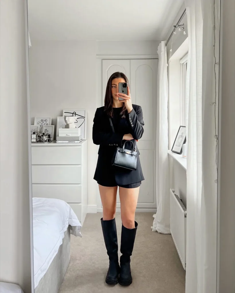 Mirror selfie of a petite woman in an all-black monochrome outfit with a longline blazer, black mini shorts, chunky lug-sole knee-high boots, and a small black structured top-handle bag