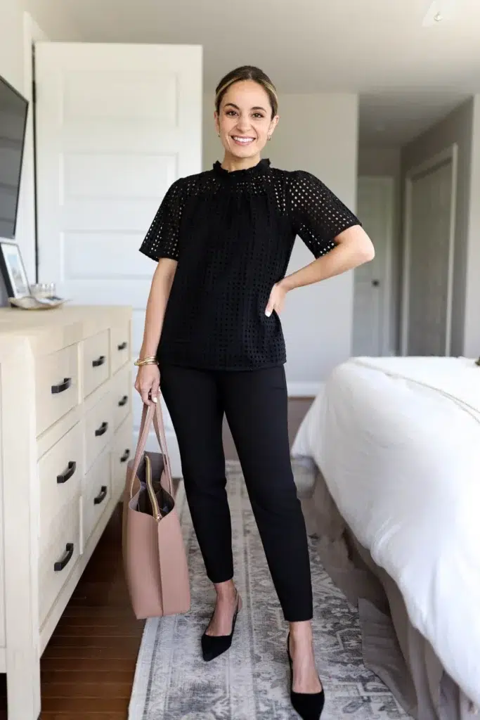 Petite woman wearing a black eyelet cutwork flutter-sleeve top with a ruffled mock neckline and a loose boxy fit, paired with slim black ankle trousers, black pointed-toe suede pumps, and a blush open structured tote bag, a tonal all-black low maintenance petite work outfit with textural contrast