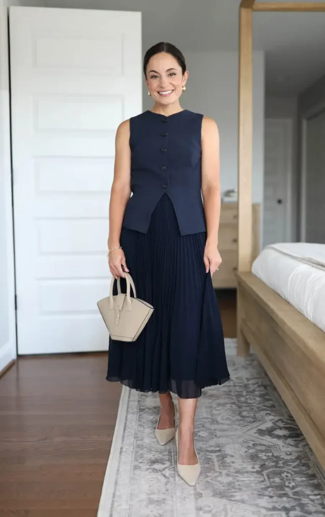 Petite woman wearing an all-navy monochrome outfit with a fitted button-front sleeveless vest and fine pleated midi skirt, paired with nude pointed-toe pumps and a small structured beige top-handle bag