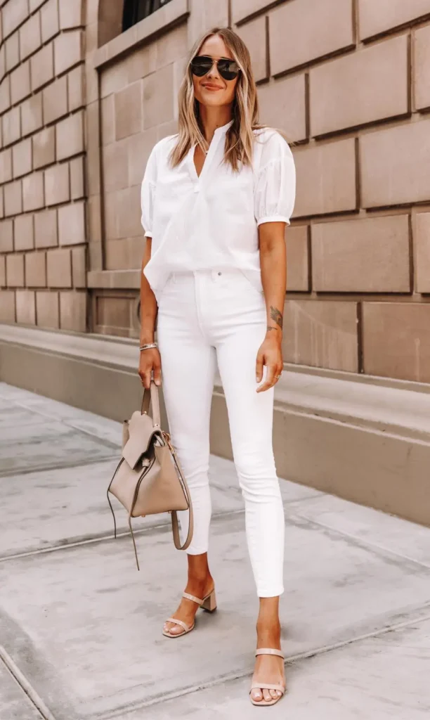 Woman in an all-white monochrome outfit featuring a puff-sleeve blouse and high-waisted white skinny jeans, accessorised with nude strappy sandals and a structured beige bag on a city street