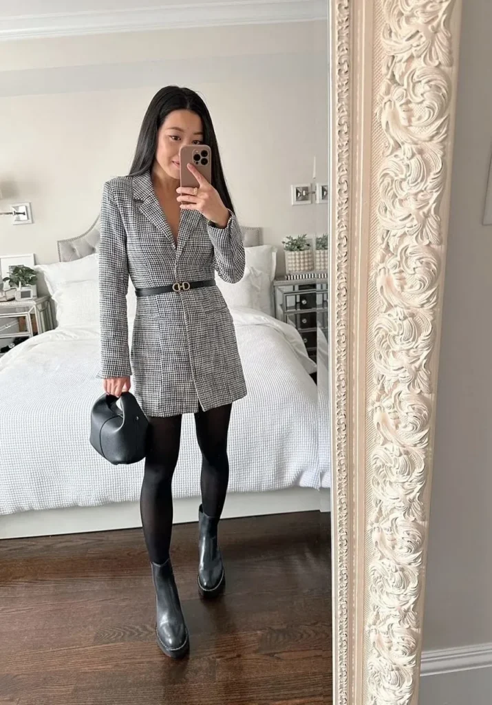 Petite woman wearing a belted houndstooth blazer dress with black opaque tights and black ankle boots, mirror selfie