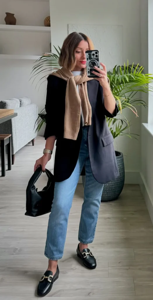 Black blazer outfit with a camel knit scarf draped over the lapels, light wash straight jeans cuffed at the ankle, black gold-hardware loafers, and a soft black hobo bag