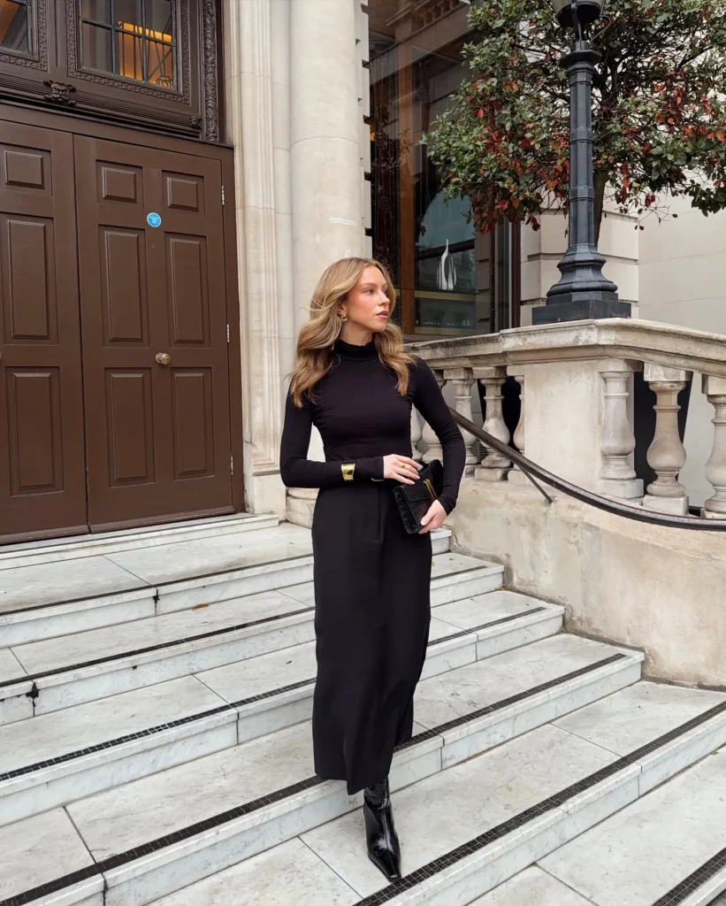 Petite woman in an all-black monochrome outfit with a fitted long-sleeve turtleneck, straight column maxi skirt, and patent pointed ankle boots, accessorised with a chunky gold cuff, gold earrings, and a black clutch