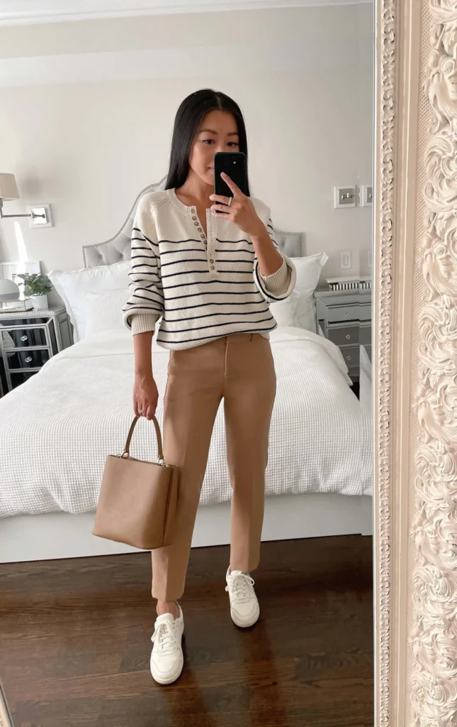 Petite woman wearing a cream and navy stripe henley knit sweater with camel ankle-length tailored trousers, white sneakers, and a tan structured top-handle bag, an effortless low maintenance petite work outfit with a French-inspired feel