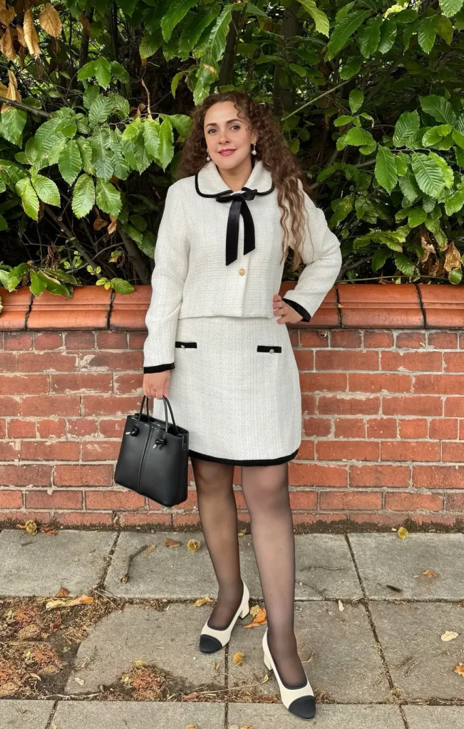 Petite woman in a cream boucle tweed co-ord with black trim detailing on the cropped jacket and mini skirt, styled with sheer black tights, two-tone cap-toe heels, a black velvet neck bow, and a black structured tote