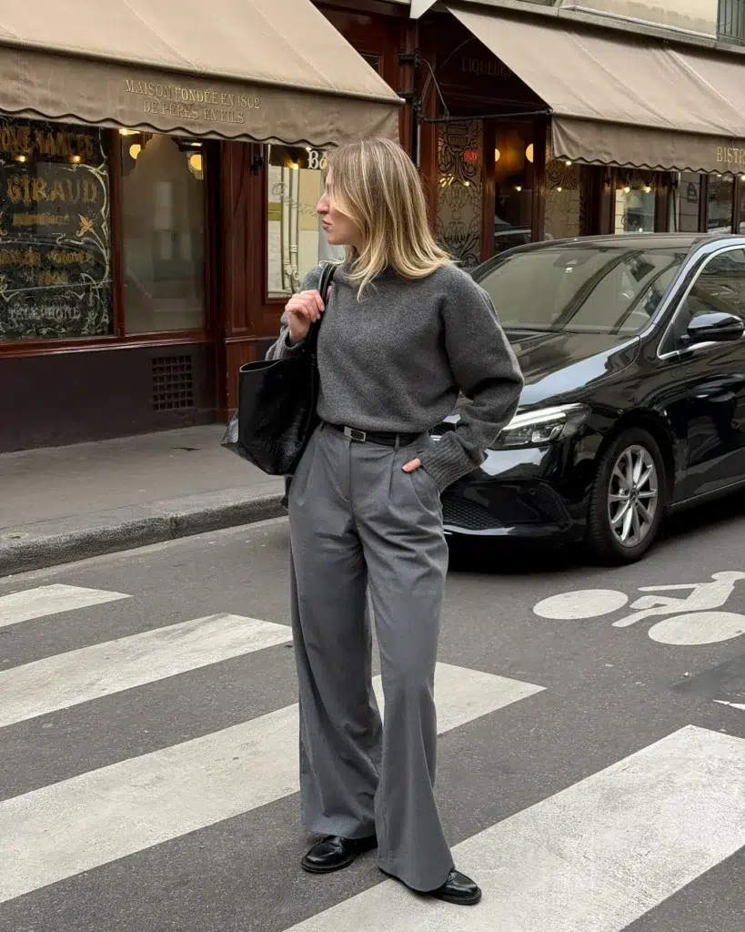 Petite woman crossing a Paris street in a grey monochrome outfit with a charcoal chunky knit sweater, mid-grey wide-leg tailored trousers, a slim black belt at the waist, black loafers, and a large black tote bag