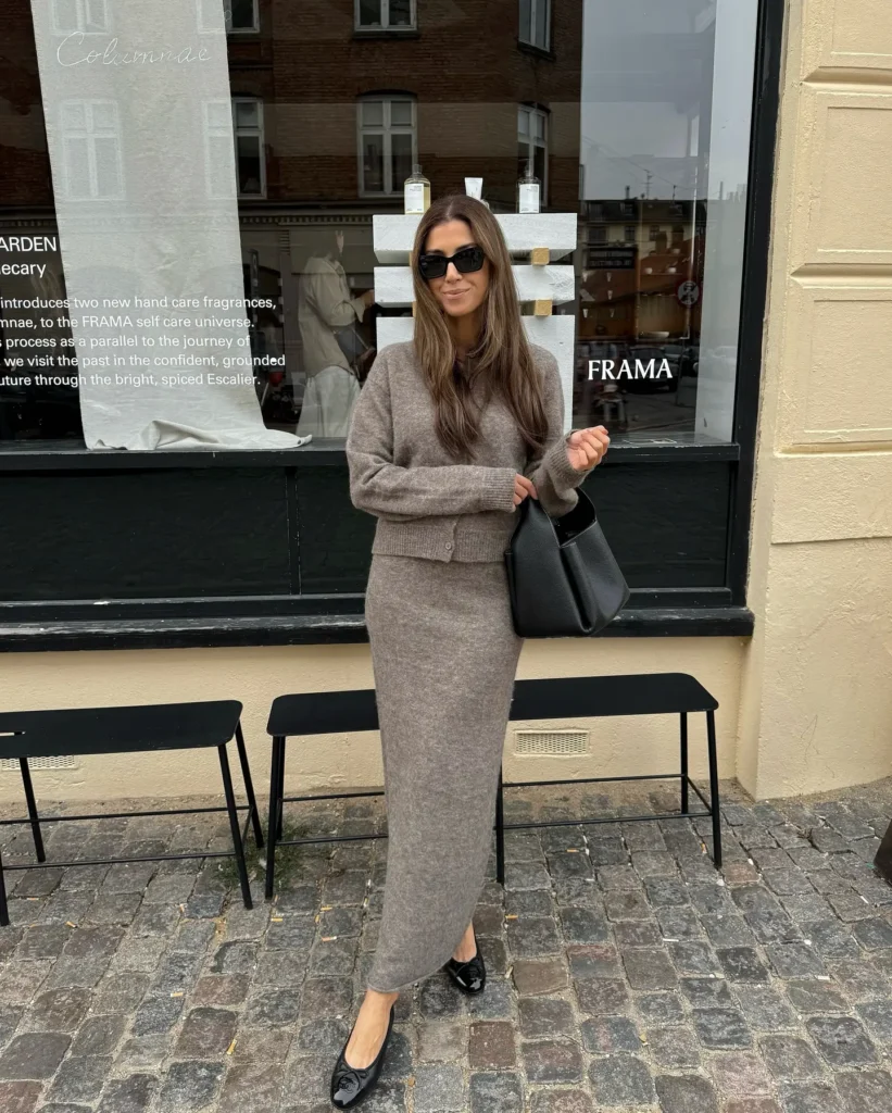Petite woman in a matching taupe heathered knit co-ord set with a cropped button-front cardigan and slim column maxi skirt, paired with black pointed ballet flats and a black structured shoulder bag