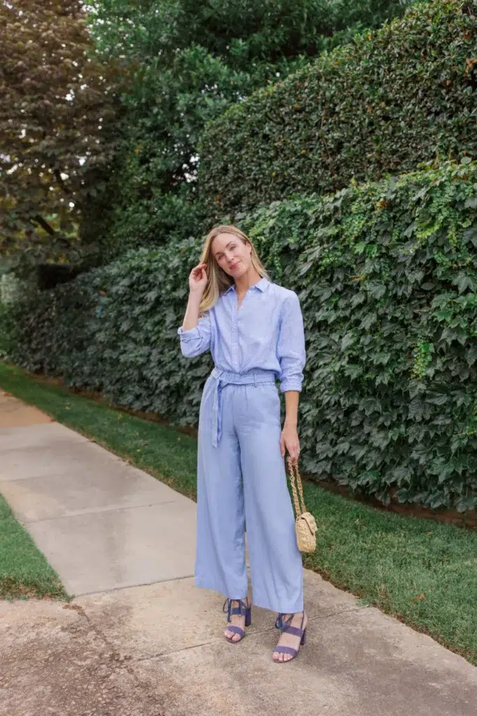 Petite woman in a periwinkle blue linen monochrome set with a collared long-sleeve shirt and cropped wide-leg trousers with a self-tie sash waist, paired with navy lace-up block heels and a small woven straw bag