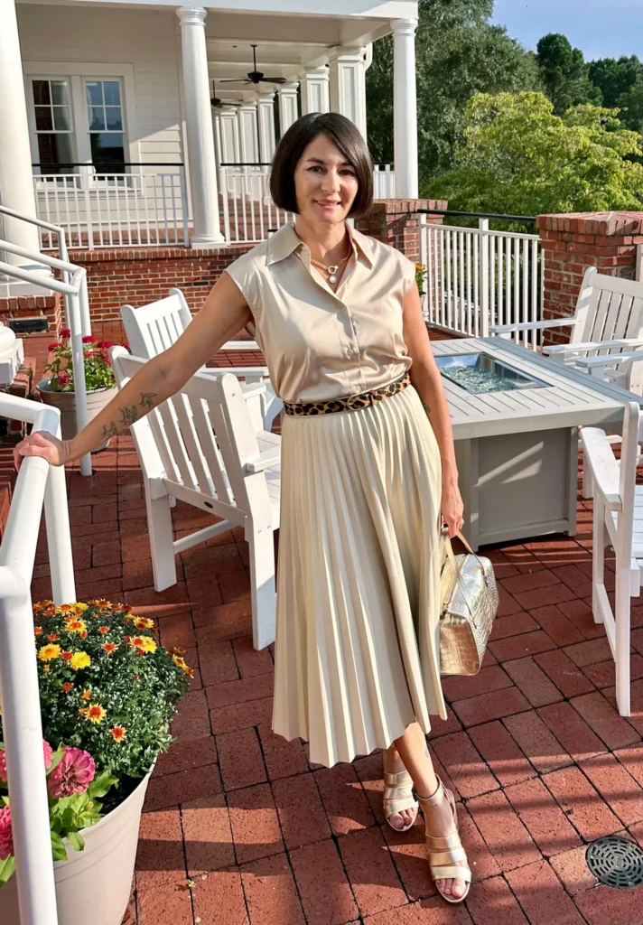 Petite woman in a sandy beige monochrome outfit with a sleeveless collared top and fine-knife pleated midi skirt, styled with a slim leopard print belt, gold block-heel sandals, and a metallic croc-embossed bag