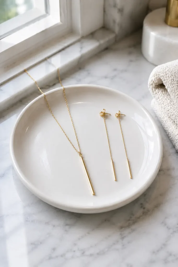 Long gold pendant necklace and thin drop earrings resting on a white ceramic dish on a marble vanity