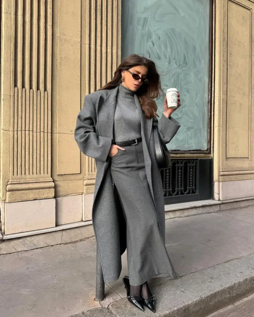 Petite woman in a full grey monochrome outfit with a fitted turtleneck, wide-leg trousers, and an oversized longline grey coat worn open, styled with a slim black belt, a black shoulder bag, and patent pointed black mules