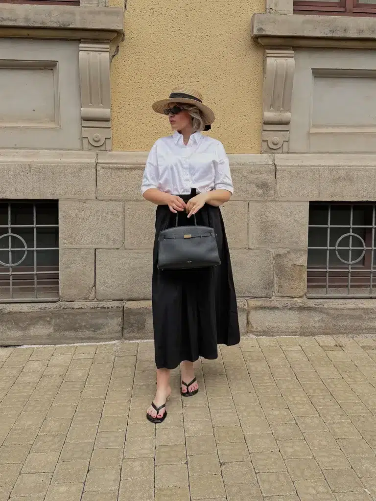 Petite woman wearing a white button-down shirt tucked into a high-waisted black full midi skirt, styled with flat black thong sandals, a straw boater hat with a black ribbon, and a structured black top-handle bag