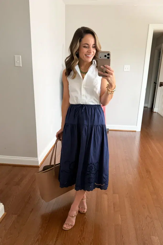 Petite woman wearing a white sleeveless button-front collared crop shirt with a navy tiered midi skirt featuring an eyelet broderie anglaise hem border, nude block-heel strappy sandals, and a tan tote bag, a feminine low maintenance petite work outfit for warmer months
