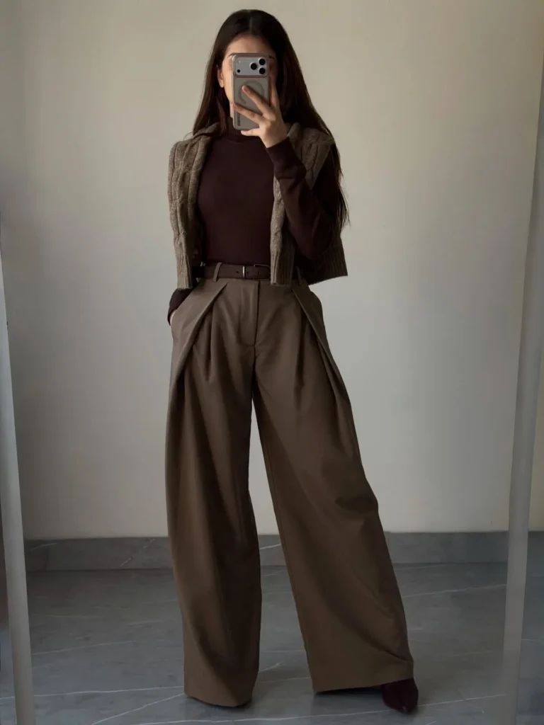 Mirror selfie of a petite monochrome brown tonal outfit with a dark chocolate ribbed turtleneck, cable-knit gilet, wide-leg pleated trousers belted at the natural waist, and deep burgundy heeled shoes