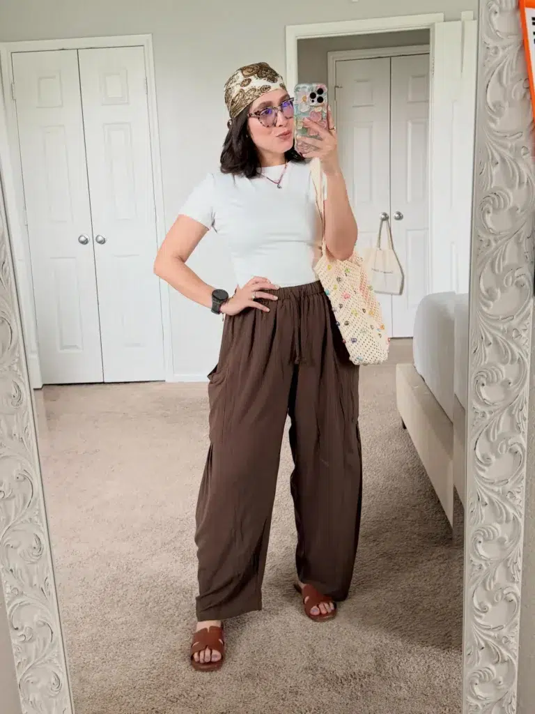 Curvy petite woman in a white ribbed fitted baby tee and chocolate brown wide-leg flowy pants with an elastic drawstring waist, wearing brown leather slide sandals and a gold paisley headscarf tied as a headband, carrying a cream crochet tote bag with colorful beaded embellishments, taking a mirror selfie in a bedroom.