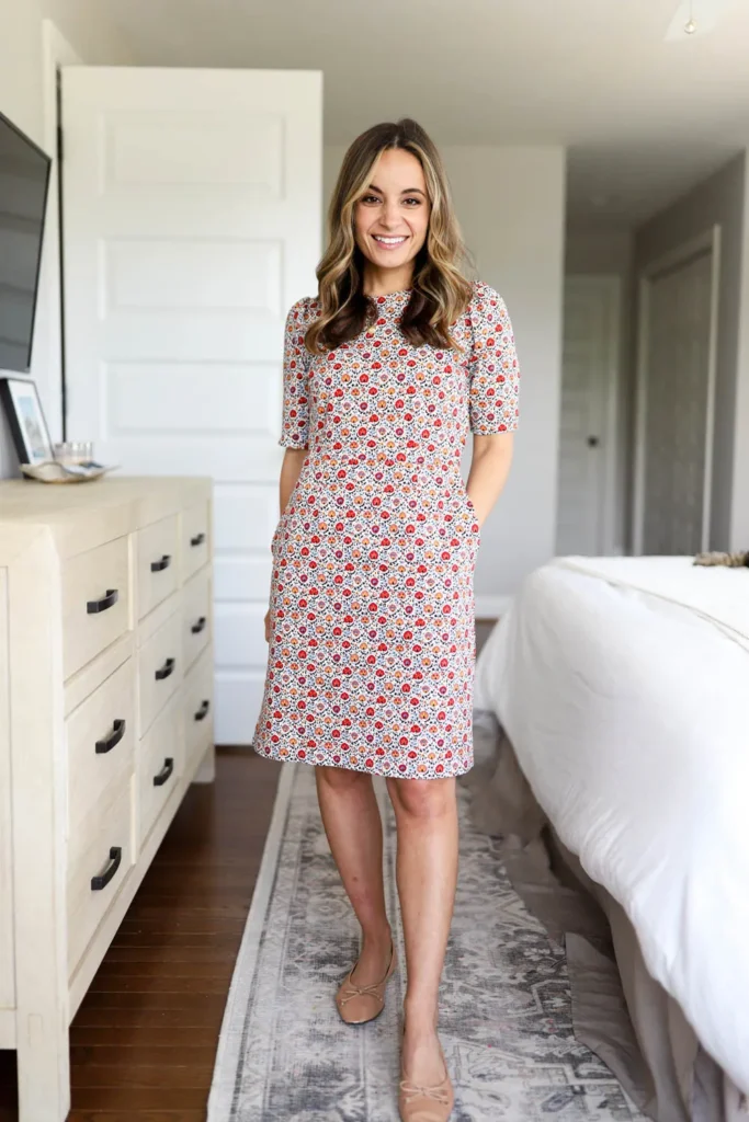 Petite woman wearing a short-sleeve red and orange floral print shift dress with side pockets and nude bow ballet flats, styled as a petite casual Friday work outfit.