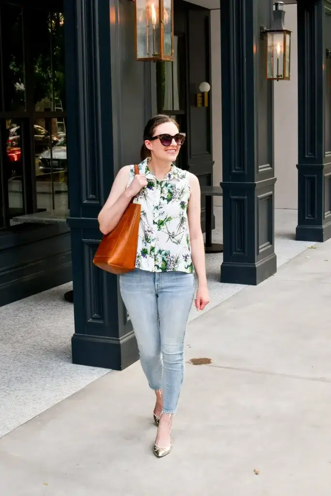 Petite woman on a city sidewalk wearing a white sleeveless botanical print button-front blouse, light wash ankle skinny jeans with a raw hem, gold metallic pointed flats, and a cognac leather shoulder tote, styled as a petite casual Friday work outfit.