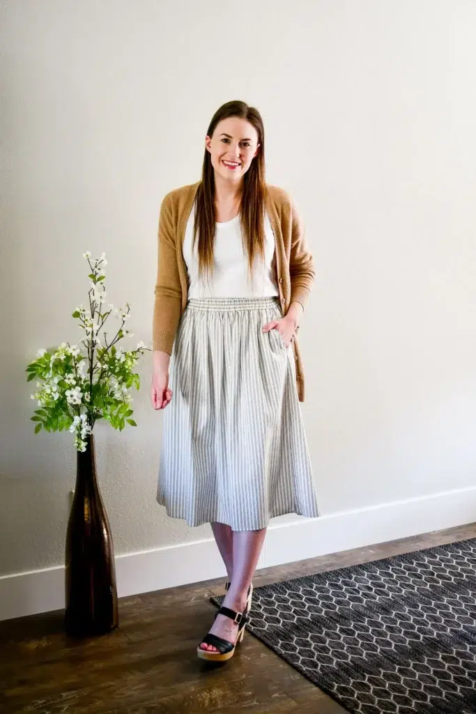 Petite woman wearing an open camel cardigan over a white tank top, a grey striped midi skirt with an elastic waist, and black platform wedge sandals, styled as a casual Friday work outfit.