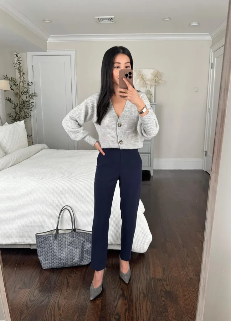 Petite woman in a mirror selfie wearing a light grey cropped ribbed cardigan with tortoiseshell buttons, high-waisted navy tapered trousers, and grey suede pointed-toe pumps, with a grey monogram tote, styled as a petite casual Friday work outfit.