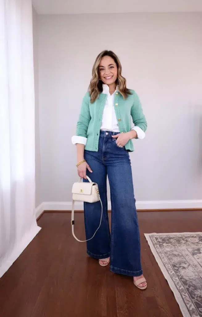 Petite woman wearing a mint green gold-button cropped cardigan over a white collared shirt, high-waisted dark indigo wide-leg jeans, nude heeled sandals, and a cream mini top-handle bag with gold hardware, styled as a petite casual Friday work outfit.