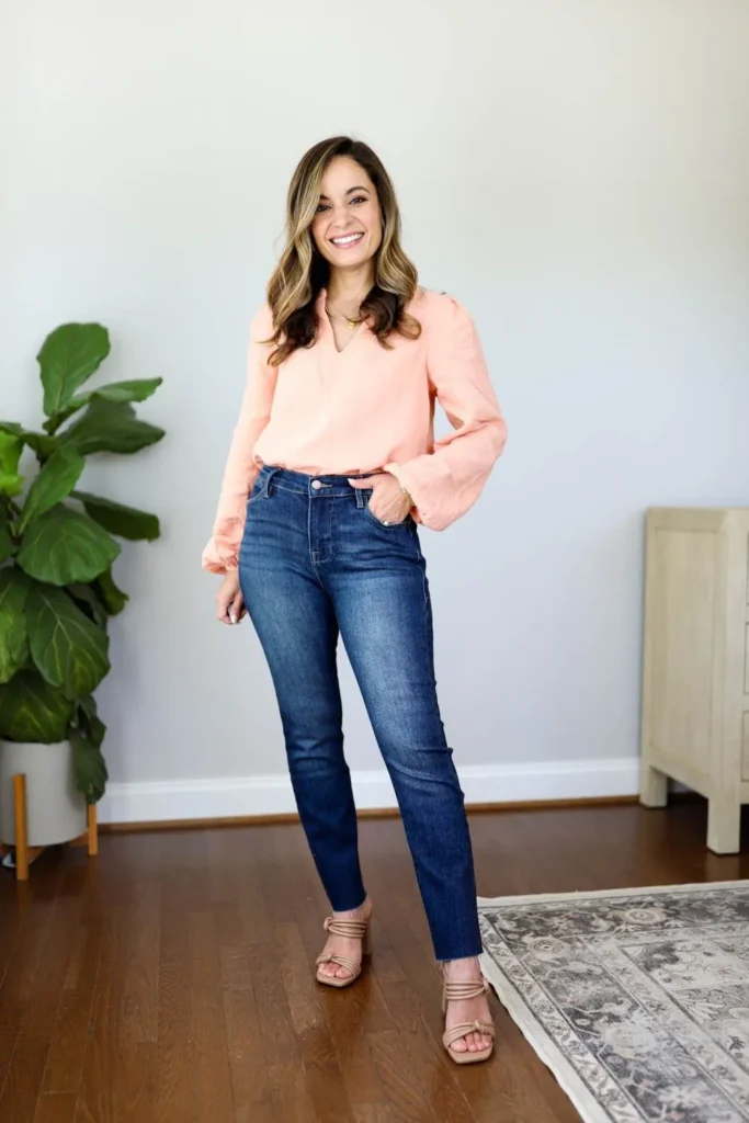 Petite woman wearing a peach long-sleeve bell-cuff V-neck blouse tucked into high-waisted dark indigo skinny jeans, with nude strappy block-heel sandals and a delicate gold necklace, styled as a petite casual Friday work outfit.