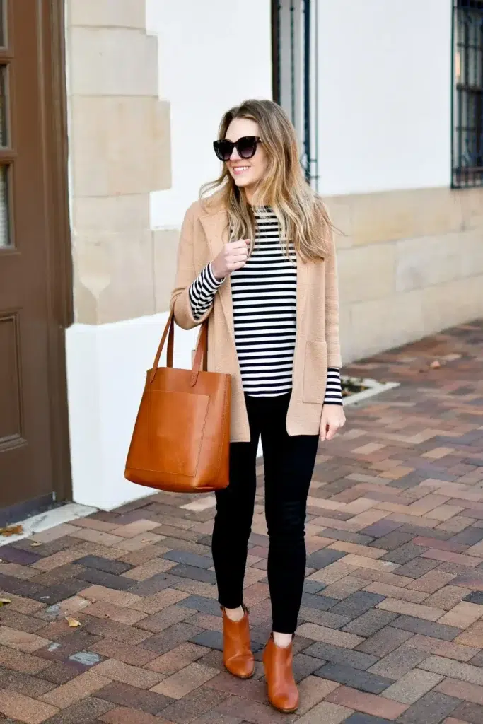 Petite woman outdoors wearing a black and white striped long-sleeve knit under an open camel longline cardigan, black skinny jeans, cognac ankle boots, and a cognac leather tote, styled as a petite casual Friday work outfit.