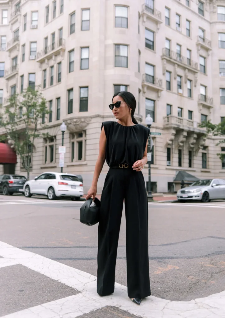 Petite woman in an all-black sleeveless structured padded-shoulder top with gathered fabric at the neckline tucked into high-rise black pleated wide-leg dress trousers cinched with a thin dark belt with a gold logo buckle, wearing black pointed-toe heels and carrying a small structured black top-handle bag, standing at a city crosswalk intersection with a grand beige stone classical building in the background.