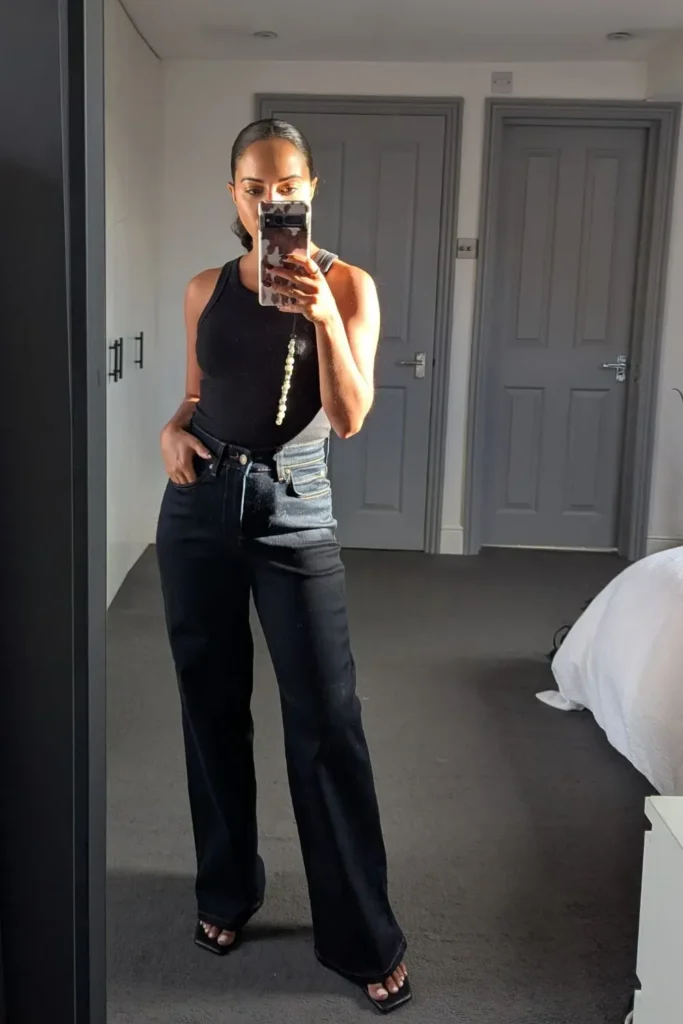 Petite woman in a fitted black sleeveless racerback bodysuit tucked into high-rise dark black wide-leg jeans with a lighter pocket lining detail visible at the hip, wearing black square-toe strappy heeled sandals and a long beaded phone charm, taking a mirror selfie in a modern bedroom with grey carpeted floors and grey panel doors visible in the background.