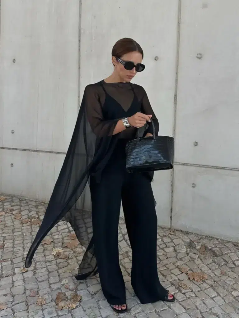 Petite woman in an all-black outfit consisting of a sheer flowing cape-style top with wide draped sleeves layered over a fitted black camisole with a visible V-neckline, paired with wide-leg black trousers reaching to the floor and black open-toe heeled mules, carrying a structured black crocodile-embossed mini tote bag with short top handles, wearing a large silver cuff bracelet and small oval dark sunglasses with a silver-detailed frame, hair pulled back in a sleek low bun, standing against a raw concrete wall on a cobblestone street with scattered dry autumn leaves on the ground.