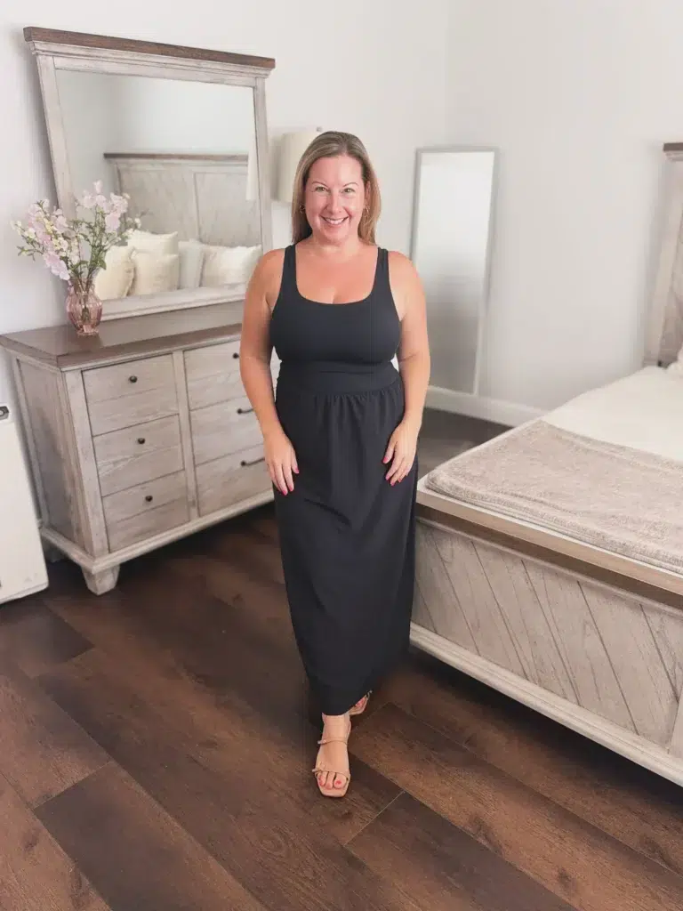 Curvy petite woman standing in a bedroom smiling, wearing a fitted all-black scoop-neck tank maxi dress with a built-in bodice, defined waist seam, and relaxed floor-length skirt, paired with tan flat strappy sandals and gold hoop earrings, with dark hardwood floors and grey wood-finish bedroom furniture in the background.