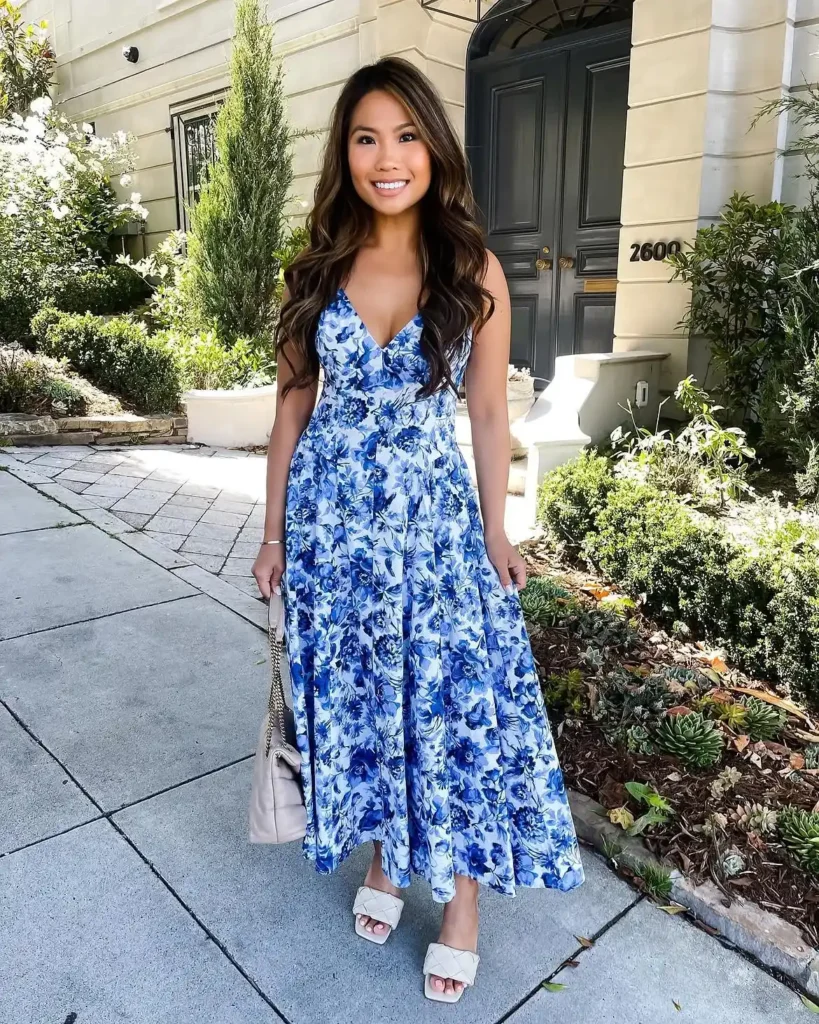 Petite woman with long wavy brown hair in an all-blue watercolor floral V-neck midi dress with a full sweeping skirt, wearing flat white quilted mule slide sandals and carrying a small beige chain-strap shoulder bag, standing on a sunny residential pavement with garden greenery and a dark arched doorway in the background.
