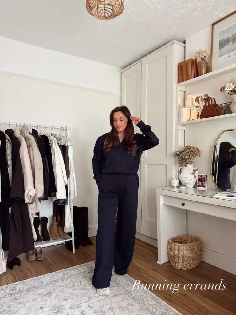 Petite woman with long dark brown hair standing in a warmly decorated home dressing room with a clothing rack, rattan pendant light, and styled shelves visible, wearing a matching all-navy coordinated set consisting of a button-front collared sweatshirt top with chest patch pockets and wide-leg tailored-style trousers with a center seam, paired with white sneakers, looking down pensively with one hand raised near her hair.