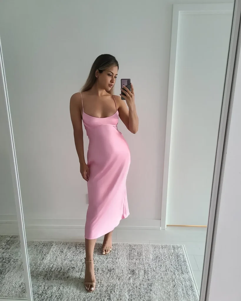 Young woman with long dark hair taking a mirror selfie wearing a bubblegum pink satin bias-cut midi slip dress with thin spaghetti straps and a low scoop neckline, paired with barely-there nude strappy heeled sandals, standing on a plush grey rug in a clean, minimal white room.