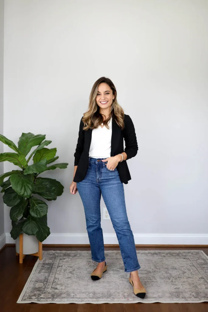 Petite woman smiling in a bright minimal white room, wearing a fitted black open blazer over a white V-neck fitted tee, medium-wash high-rise straight-leg jeans cropped at the ankle, and two-tone tan and black cap-toe pointed-toe flats, with a simple silver bracelet and loose wavy highlighted hair, standing on a vintage-style area rug beside a large fiddle leaf fig plant.
