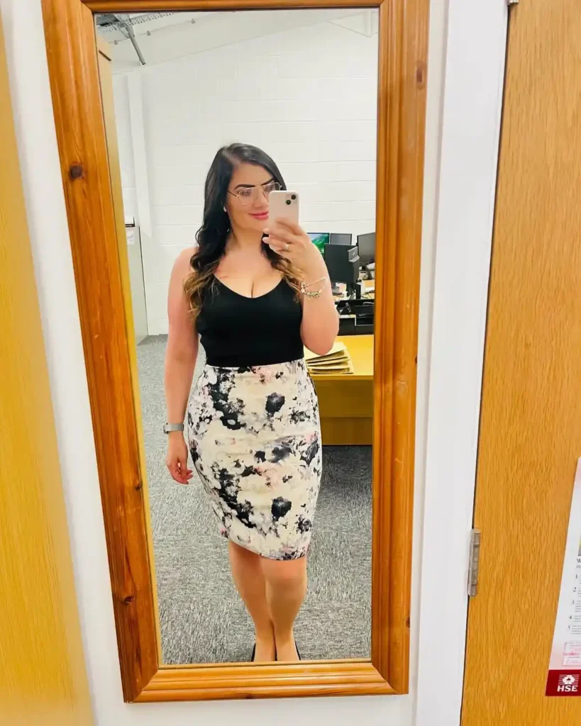 Petite woman with long dark wavy hair and glasses in a black fitted scoop-neck camisole tucked into a high-waisted fitted abstract watercolor floral print pencil skirt in cream, blush, and black, taking a mirror selfie in an office setting.