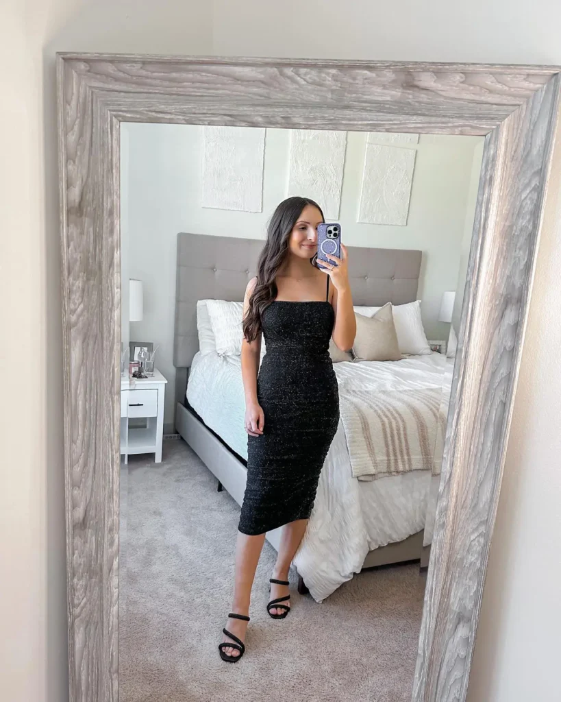 Slim petite brunette woman with long dark wavy hair taking a mirror selfie in a bedroom, wearing a black glitter-textured ruched bodycon midi dress with a square neckline and thin spaghetti straps, paired with simple black double-strap heeled sandals and a delicate gold necklace, reflected in a large grey distressed wood-framed floor mirror with a neutral beige tufted upholstered bed and white bedding visible in the background.