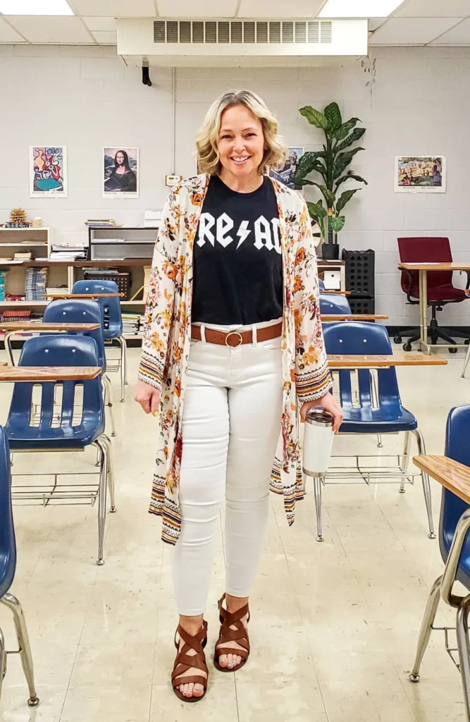 Curvy petite woman standing in a classroom wearing a black graphic tee tucked into white skinny jeans with a brown circle-buckle belt, layered under a long open floral kimono duster in cream with rust and orange blooms and a border print at the cuffs, paired with brown strappy gladiator sandals, holding a white insulated tumbler.
