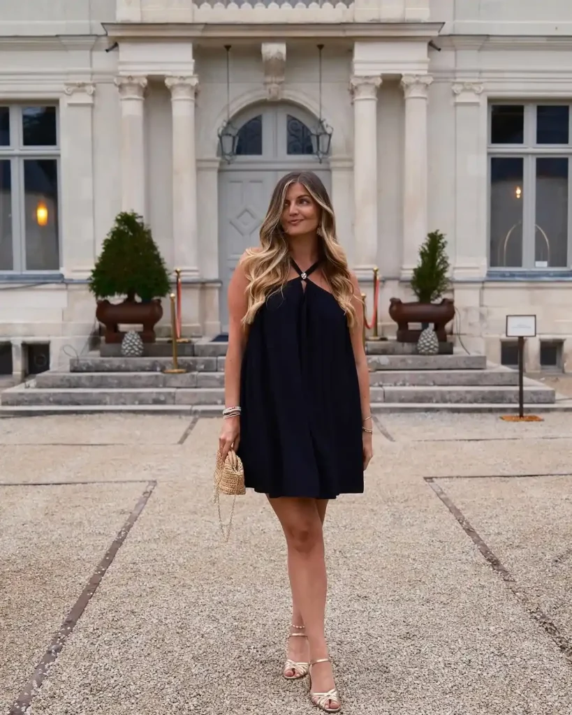 Petite woman in a black flowing halter trapeze mini dress with soft gathered draping at the chest and a small jewel clasp detail at the halter neckline, wearing gold strappy heeled sandals with ankle straps and carrying a tiny woven raffia evening bag, with stacked bracelets on both wrists and long wavy hair worn down, standing on a gravel courtyard in front of a grand classical stone building with columns at dusk.