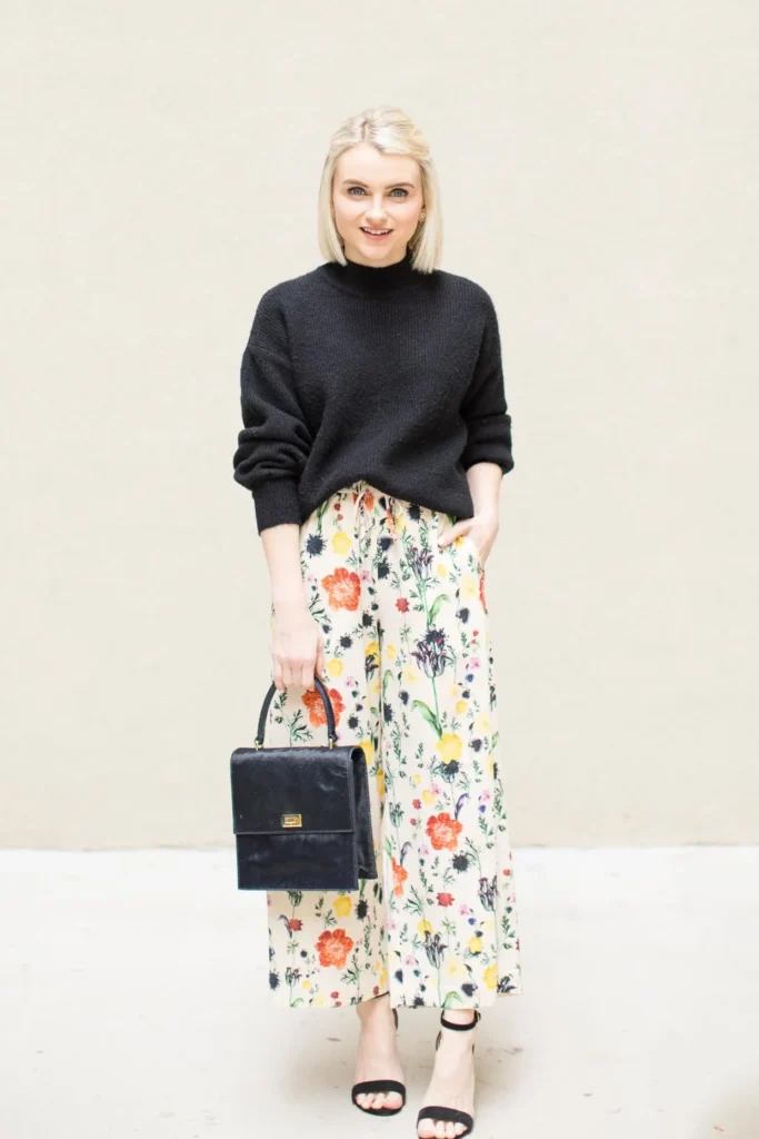Petite woman in an oversized black chunky mock-neck knit sweater paired with a cream full-length floral maxi skirt printed with botanical orange, yellow, pink and purple flowers, carrying a structured black box top-handle bag with gold hardware, wearing black strappy heeled sandals, standing against a plain warm beige wall.