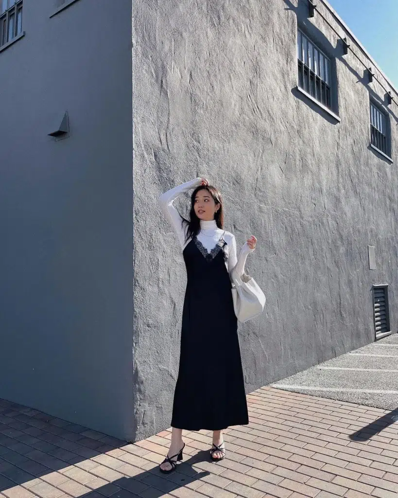 Petite East Asian woman with long dark hair standing against a textured grey stucco wall on a brick sidewalk in bright sunlight, wearing a black satin midi slip dress with delicate black lace trim at the V-neckline layered over a slim white long-sleeve fitted turtleneck, paired with thin-strap black heeled strappy sandals and a soft cream hobo shoulder bag, one hand raised to her hair while she glances to the side.