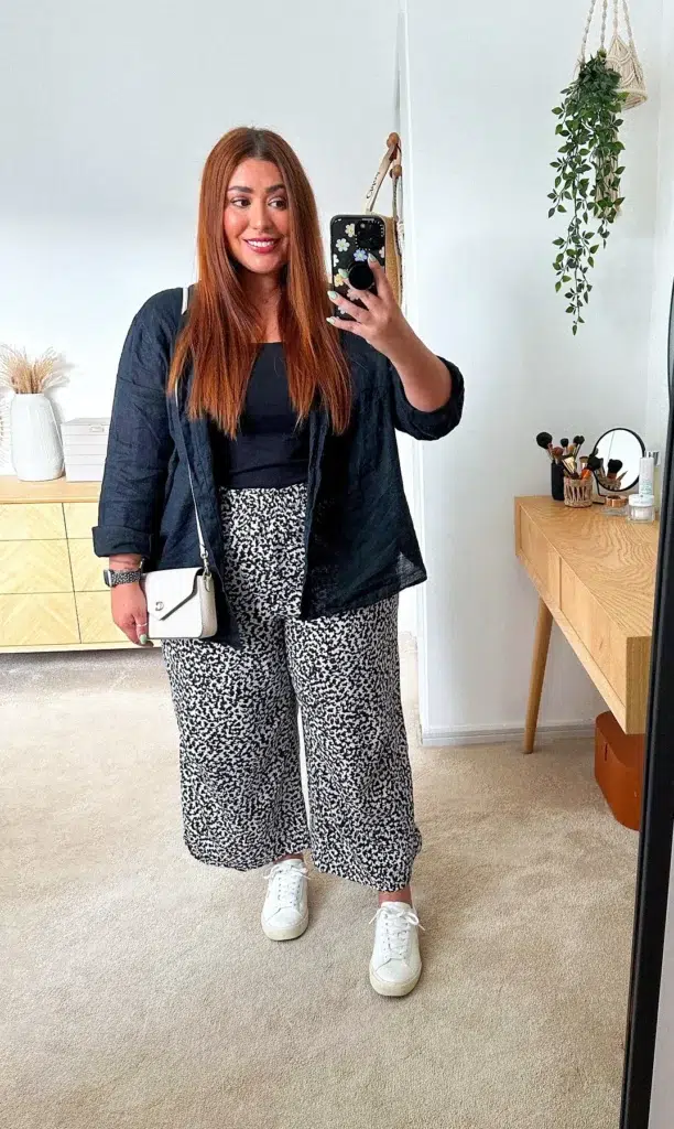 Woman with long straight auburn hair taking a mirror selfie in a modern bright bedroom, wearing a dark navy open linen-texture overshirt over a fitted black camisole, black and white ditsy print wide-leg culottes cropped above the ankle, white low-profile leather sneakers, and a small white crossbody bag with gold hardware, with a hanging trailing plant and wooden vanity table visible in the background.