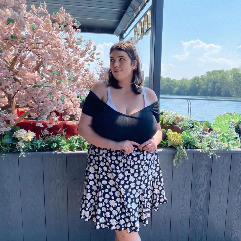 Petite plus-size woman with medium-length dark brunette hair worn half-up, wearing a black off-shoulder bardot top with a visible white cami or bralette strap at the shoulder, paired with a knee-length black flared skirt covered in scattered white and yellow daisy flowers, standing on a modern rooftop terrace with gray wood paneling, mixed flower planters, a decorative cherry blossom tree in the background, and a river and blue sky visible beyond the railing.