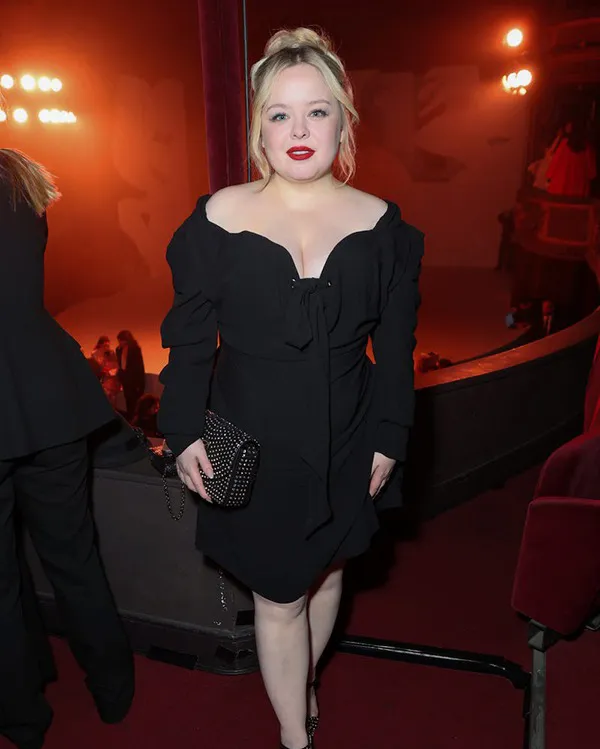 Petite woman in a black off-shoulder long-sleeve mini dress with a deep sweetheart neckline, structured puffed sleeves, and a bow tie detail at the front waist, wearing dark pointed heels and holding a black studded chain clutch, standing at an indoor evening event with warm red ambient lighting.