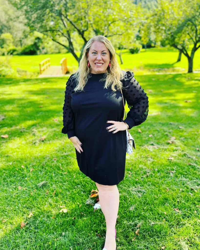 Curvy petite woman standing on a green lawn with trees in the background wearing a black mock-neck mini dress with long sheer organza sleeves covered in three-dimensional pom pom dot texture, styled with hot pink drop earrings, pink heeled shoes, and a small colorful monogram print bag worn crossbody, with long wavy blonde hair worn loose.