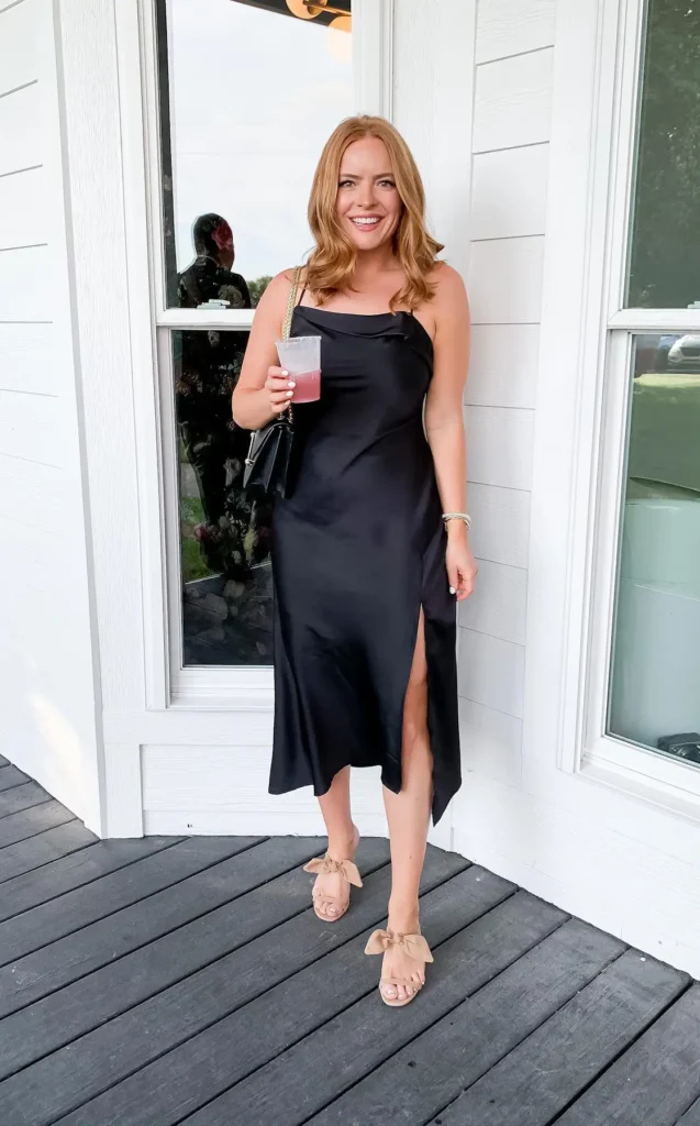 Woman wearing a black satin slip midi dress with a cowl neckline, spaghetti straps, and a side slit, paired with nude bow flat sandals and a black chain-strap shoulder bag.