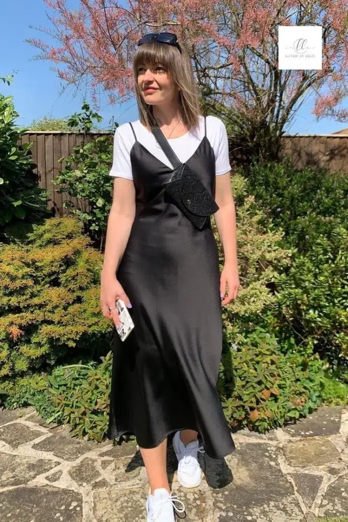 Petite woman in a black satin bias-cut midi slip dress layered over a white crew-neck t-shirt, wearing white chunky lace-up sneakers and a black beaded crossbody belt bag, standing outdoors in a sunny garden with pink flowering trees in the background.