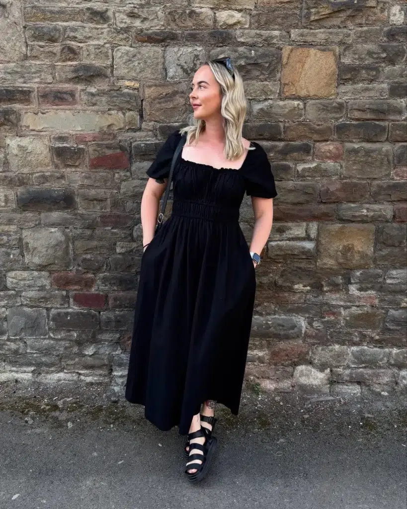 Petite woman standing against a stone wall, wearing an all-black cotton milkmaid maxi dress with a square neckline, short puff sleeves, and a shirred bodice, chunky black strappy sandals, sunglasses pushed up on her head, and a black crossbody bag.