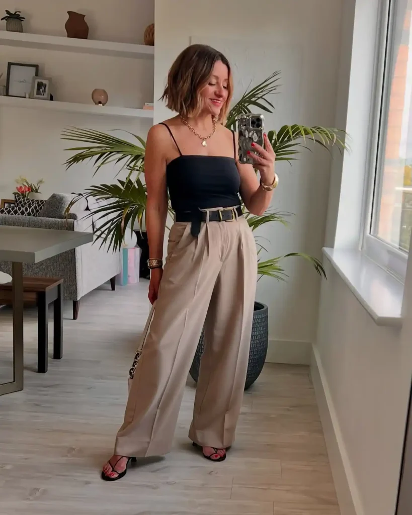 Petite woman in a fitted black square-neck spaghetti strap cami tucked into high-rise sand-toned pleated wide-leg tailored trousers with a wide black leather belt, wearing strappy black kitten heel sandals and carrying a gold chain-strap bag, with a chunky gold chain pendant necklace and a gold cuff bracelet, taking a mirror selfie in a bright modern living room with a large palm plant behind her.