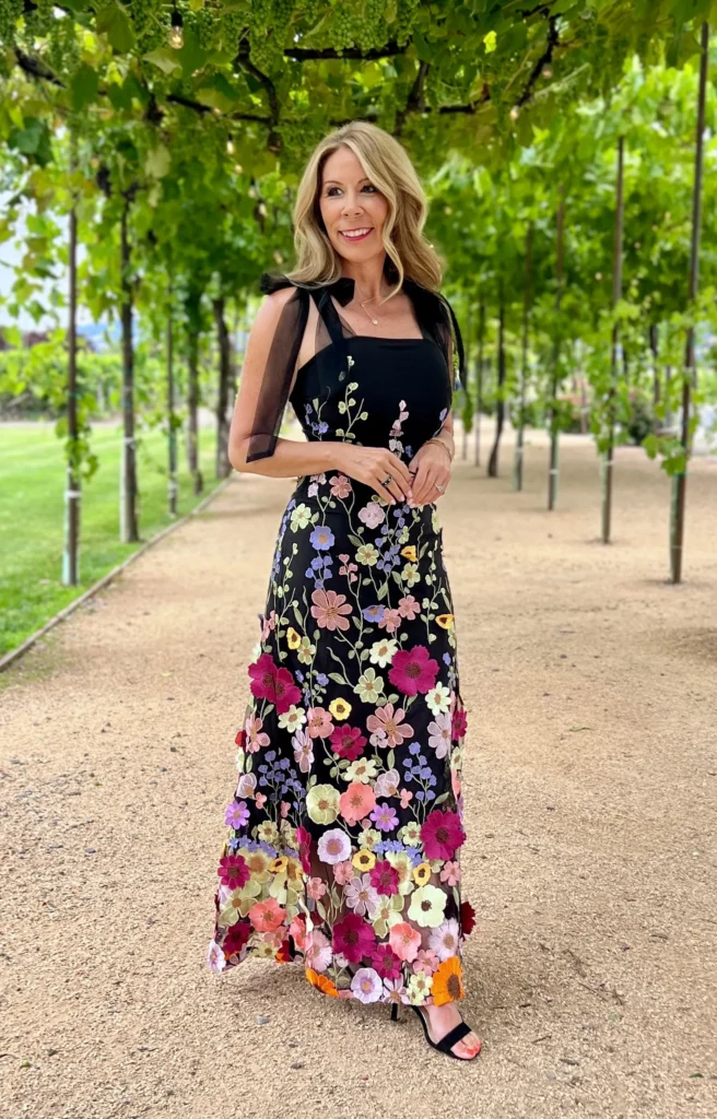 Woman wearing a black maxi dress with a fitted square-neck bodice, black organza bow shoulder straps, and a full A-line skirt covered in dense multi-colored 3D floral appliqués in pink, purple, lavender, coral, and yellow, paired with black strappy heeled sandals at a vineyard wedding.